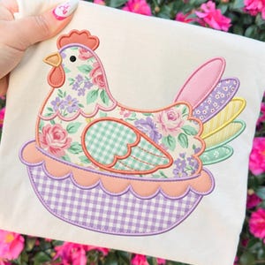May include: A cream-colored fabric square featuring a colorful appliqué of a hen. The hen is adorned with floral patterns in pink, purple, and green, and sits in a scalloped basket with a gingham base.