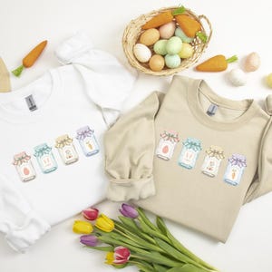 May include: Two crewneck sweatshirts, one white and one tan, each with a colorful jar graphic. The jars have pastel lids and contain Easter-themed designs. A basket of eggs and carrots, a straw hat, and tulips complete the spring theme.