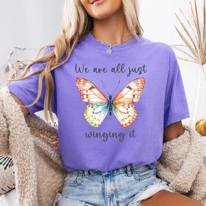May include: A lilac-colored t-shirt featuring a watercolor butterfly with orange, yellow, green, and blue wings. The text "We are all just winging it" is printed below the butterfly. The shirt is part of a clothing collection.
