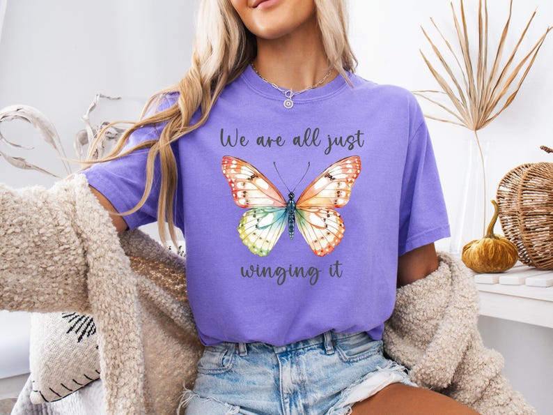 May include: A lilac-colored t-shirt featuring a watercolor butterfly with orange, yellow, green, and blue wings. The text "We are all just winging it" is printed below the butterfly. The shirt is part of a clothing collection.