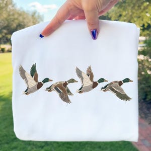 May include: A white sweatshirt featuring an embroidered design of four flying ducks. The ducks are depicted with detailed wings in shades of brown, gray, and green, with green heads. The sweatshirt is held up by a hand.