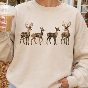 May include: A beige crewneck sweatshirt featuring a graphic print of four deer standing in a row. Each deer has a different patterned body, including camouflage and plaid designs in shades of brown, green, and tan, set against the light fabric.