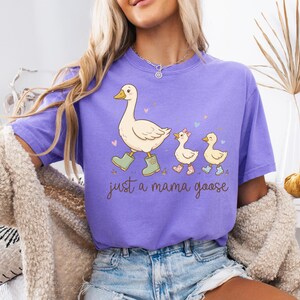 May include: A lilac-colored t-shirt featuring a cartoon goose leading two goslings. The mama goose wears green boots, and the goslings wear pink and blue boots. The text "just a mama goose" is written in a script font.
