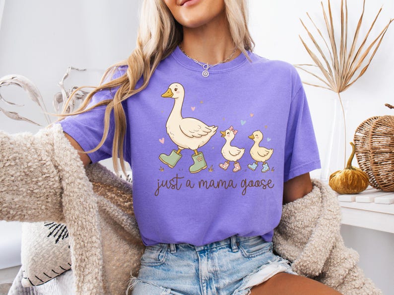 May include: A lilac-colored t-shirt featuring a cartoon goose leading two goslings. The mama goose wears green boots, and the goslings wear pink and blue boots. The text "just a mama goose" is written in a script font.