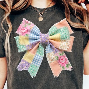 Pu&ograve; includere: T-shirt grigio scuro con un grande fiocco patchwork. Il fiocco &egrave; realizzato con vari tessuti dai colori pastello, tra cui motivi floreali, a quadretti e tinta unita. Il fiocco &egrave; il punto focale della maglietta.