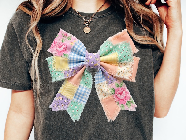 Pu&ograve; includere: T-shirt grigio scuro con un grande fiocco patchwork. Il fiocco &egrave; realizzato con vari tessuti dai colori pastello, tra cui motivi floreali, a quadretti e tinta unita. Il fiocco &egrave; il punto focale della maglietta.