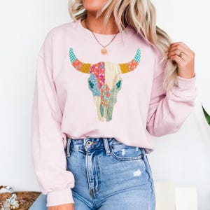 May include: A light pink sweatshirt featuring a colorful bull skull design. The skull is decorated with floral and plaid patterns in shades of pink, blue, yellow, and green. The model is wearing blue jeans.