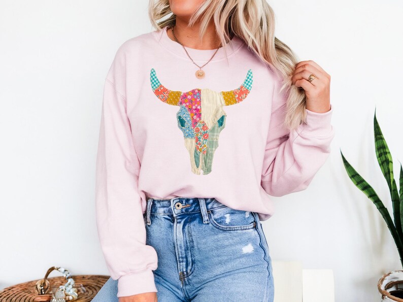 May include: A light pink sweatshirt featuring a colorful bull skull design. The skull is decorated with floral and plaid patterns in shades of pink, blue, yellow, and green. The model is wearing blue jeans.