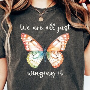 May include: Dark gray t-shirt featuring a colorful butterfly graphic with the text "We are all just winging it." The butterfly design has orange, red, yellow, green, and blue wings. The shirt is a casual style.