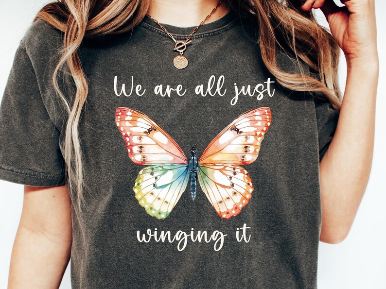 May include: Dark gray t-shirt featuring a colorful butterfly graphic with the text "We are all just winging it." The butterfly design has orange, red, yellow, green, and blue wings. The shirt is a casual style.