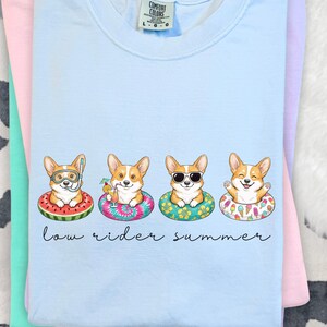 May include: Pale blue t-shirt with a graphic of four corgi dogs in inflatable rings. Each corgi has a different accessory. The text "low rider summer" is written below the corgis. The shirt is folded with other pastel-coloured shirts.