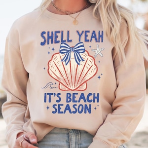 May include: A tan sweatshirt with the words "SHELL YEAH" and "IT'S BEACH SEASON" in blue text. A seashell graphic with a blue bow and a starfish are in the center. The sweatshirt is a casual beach-themed apparel item.
