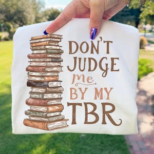 Może przedstawiać: Biała koszulka z ilustracją stosu książek w odcieniach brązu i beżu. Koszulka ma napis "DON'T JUDGE me BY MY TBR" w brązowych literach. Projekt jest umieszczony na środku przodu koszulki.