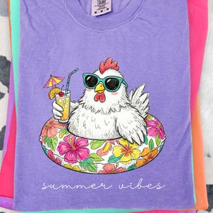 May include: A lilac-colored t-shirt featuring a cartoon chicken wearing sunglasses and floating in a floral inner tube, holding a tropical drink. The words "summer vibes" are written in cursive. Other folded shirts are in the background.