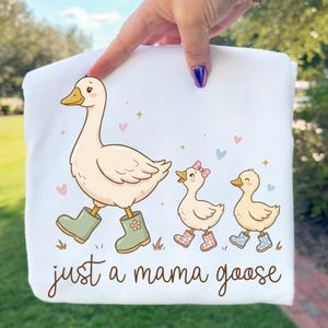 May include: White sweatshirt featuring a cartoon goose wearing green boots, leading two ducklings in patterned boots. The text "just a mama goose" is written in a script font. Pastel hearts and stars decorate the design.