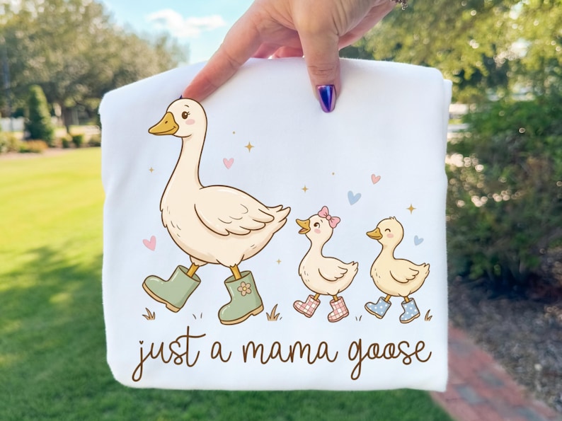May include: White sweatshirt featuring a cartoon goose wearing green boots, leading two ducklings in patterned boots. The text "just a mama goose" is written in a script font. Pastel hearts and stars decorate the design.
