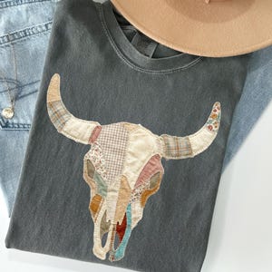 Op de afbeelding: Een donkergrijs t-shirt met een patchwork stierenkopontwerp in verschillende kleuren en patronen. Het shirt wordt gecombineerd met lichtblauwe jeans en een bruine hoed. Een zilveren ketting is zichtbaar.