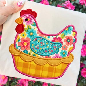 May include: A decorative fabric applique featuring a colorful hen design. The hen is adorned with floral patterns in pink, blue, and yellow, set against a cream background. It is placed in a yellow plaid basket, outlined with pink and gold stitching.