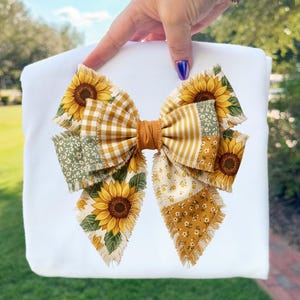 May include: A large, decorative bow featuring a patchwork design with sunflowers, gingham, and floral patterns in shades of yellow, green, and white. The bow has a frayed edge and a brown center, set against a white background.