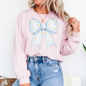 May include: A light pink sweatshirt featuring a large bow design. The bow has a blue and white gingham pattern with white daisy accents. The sweatshirt is a casual style.