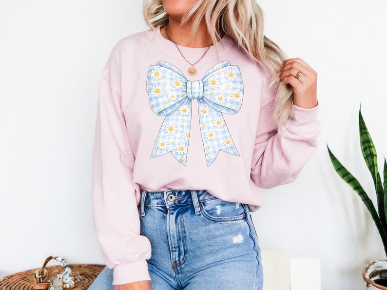 May include: A light pink sweatshirt featuring a large bow design. The bow has a blue and white gingham pattern with white daisy accents. The sweatshirt is a casual style.