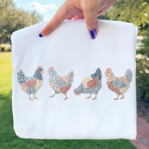 May include: White sweatshirt featuring four embroidered chickens with patchwork-style details in blue, pink, and tan. The chickens are in various poses, adding a whimsical touch to the garment. The design is centered on the front of the sweatshirt.