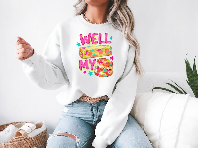 May include: A white long-sleeve crewneck sweatshirt featuring a graphic print with the text WELL MY in bright pink letters. The design includes a rectangular block of butter and two stacked biscuits, both decorated with colourful confetti sprinkles.