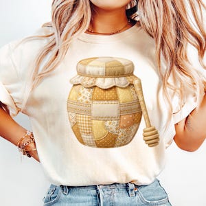 May include: A beige t-shirt with a patchwork honey pot design and a matching dipper. The honey pot is made of various fabrics in shades of yellow and beige, with floral and gingham patterns. The dipper is wooden.
