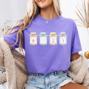 May include: A purple t-shirt featuring four jars with Easter-themed designs. Each jar has a different illustration: an egg, bunny ears, flowers, and a carrot. The jars are decorated with colorful lids and bows. The shirt is worn by a person.