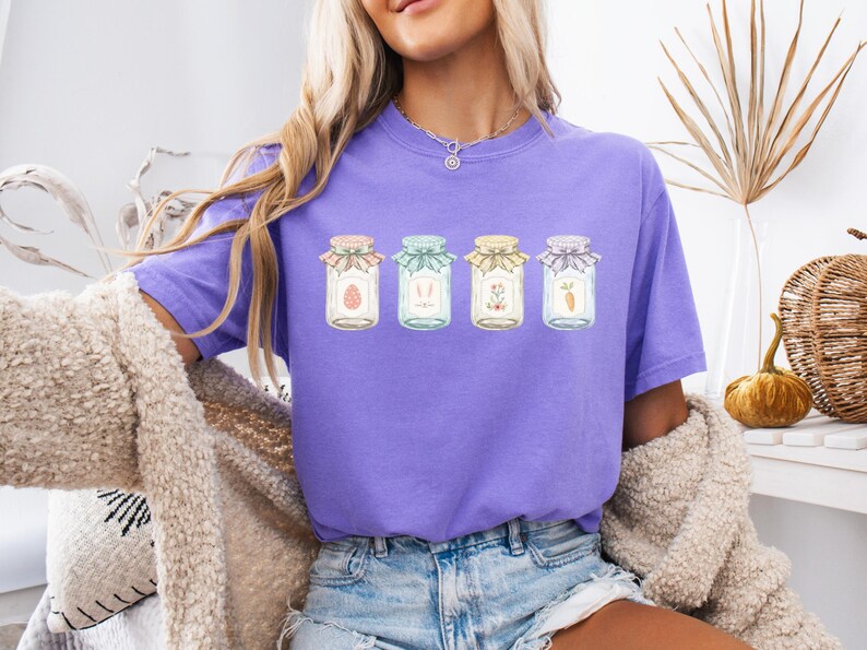 May include: A purple t-shirt featuring four jars with Easter-themed designs. Each jar has a different illustration: an egg, bunny ears, flowers, and a carrot. The jars are decorated with colorful lids and bows. The shirt is worn by a person.