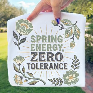 Peut inclure: T-shirt blanc avec l'inscription "SPRING ENERGY, ZERO TOLERANCE" en vert et marron. Le t-shirt présente des motifs floraux verts et beiges. Le t-shirt est tenu par une personne.