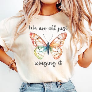 May include: Cream-colored t-shirt featuring a colorful butterfly illustration with text that reads "We are all just winging it". The butterfly has orange, red, blue, and green wings.
