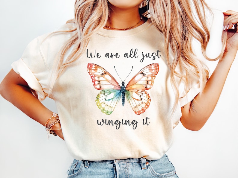 May include: Cream-colored t-shirt featuring a colorful butterfly illustration with text that reads "We are all just winging it". The butterfly has orange, red, blue, and green wings.