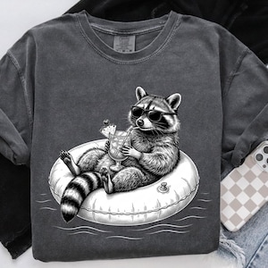 May include: Dark gray t-shirt featuring a black and white illustration of a raccoon wearing sunglasses and relaxing on an inflatable ring, holding a cocktail. The raccoon has a striped tail. The shirt is paired with jeans and a phone case.