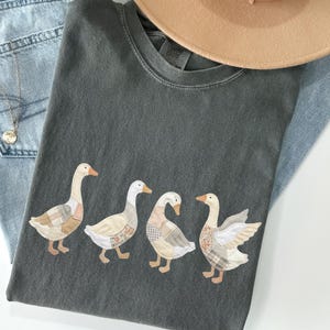 May include: Dark gray t-shirt featuring four geese appliqués with patchwork-style wings and bodies. The shirt is paired with light blue denim jeans, a gold necklace, and a tan hat. A casual, everyday outfit.