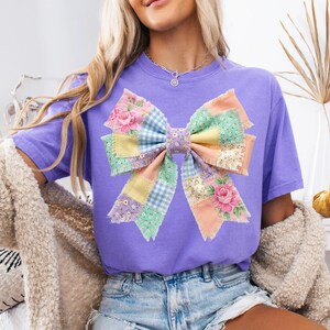 Pu&ograve; includere: Una maglietta lilla con un grande design a fiocco patchwork. Il fiocco incorpora vari motivi floreali e a quadri in colori pastello, tra cui rosa, giallo e blu. La maglietta &egrave; uno stile casual, adatto per l'uso quotidiano.