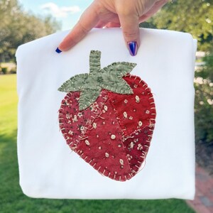 Può includere: Articolo in tessuto bianco con un grande design a fragola applicato. La fragola è rossa con accenti floreali bianchi e un gambo e foglie verdi. Il disegno è cucito sul tessuto.