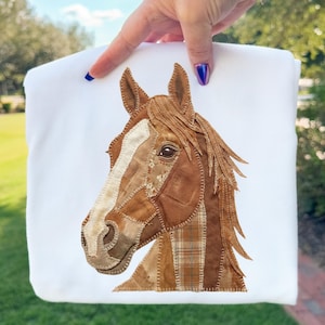 May include: White sweatshirt featuring a detailed horse head design. The horse's head is crafted from various shades of brown fabric, with a white stripe down the face. The mane and ears are also brown, creating a textured, patchwork appearance.