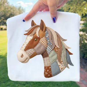 May include: A white fabric item with a patchwork horse head design. The horse head is made from various fabrics in shades of brown, beige, and blue, with a detailed mane and eye. The design is held up by a hand.