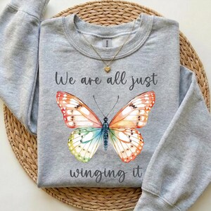 May include: A light gray sweatshirt featuring a colorful butterfly design with orange, red, green, and blue wings. The text "We are all just winging it" is printed below the butterfly. A gold necklace with a heart pendant is visible.