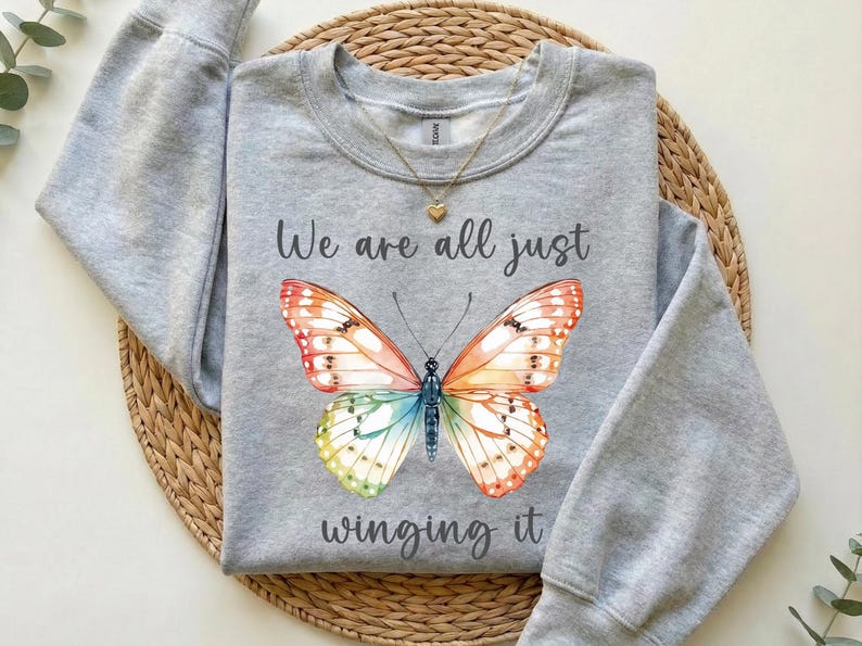 May include: A light gray sweatshirt featuring a colorful butterfly design with orange, red, green, and blue wings. The text "We are all just winging it" is printed below the butterfly. A gold necklace with a heart pendant is visible.