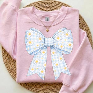 May include: A light pink sweatshirt with a large bow design. The bow features a blue and white gingham pattern with white daisies and yellow centers. A gold necklace with a heart pendant is visible.