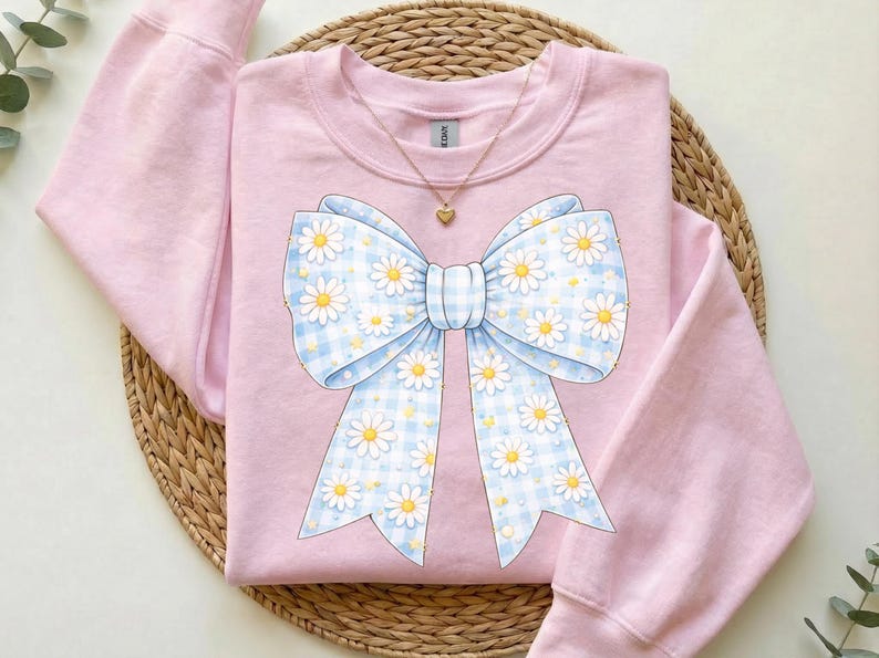 May include: A light pink sweatshirt with a large bow design. The bow features a blue and white gingham pattern with white daisies and yellow centers. A gold necklace with a heart pendant is visible.