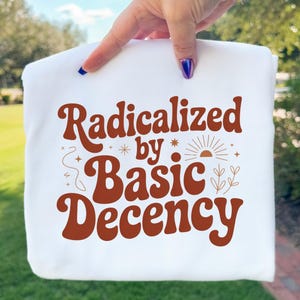 May include: White sweatshirt with the phrase "Radicalized by Basic Decency" in a retro brown font. The design includes a sun, stars, and floral elements. The sweatshirt is held up against a background of green grass and trees.