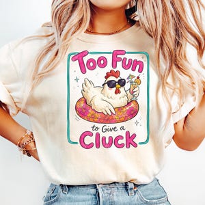 May include: Cream-colored t-shirt with a cartoon chicken wearing sunglasses and floating on a pink and yellow inflatable ring. The text reads "Too Fun to Give a Cluck" in pink and green.
