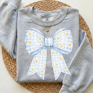 May include: A light gray sweatshirt featuring a large bow design. The bow is light blue with a gingham pattern and white daisies. A gold necklace with a heart pendant is visible.