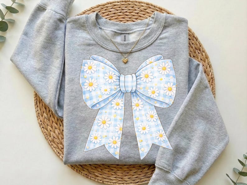 May include: A light gray sweatshirt featuring a large bow design. The bow is light blue with a gingham pattern and white daisies. A gold necklace with a heart pendant is visible.