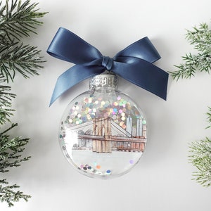 May include: A clear glass Christmas ornament with a blue ribbon bow. The ornament features a hand-drawn illustration of the Brooklyn Bridge with glitter inside.