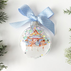 May include: A clear glass Christmas ornament with a blue ribbon and white polka dots. The ornament features a scene of ballerinas in a circle with a larger ballerina in the center.