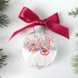 Op de afbeelding: Een transparante glazen kerstbal met een rode strik en twee ballerinafiguren erin. De bal is gevuld met kleurrijk confetti en snoepstokken.